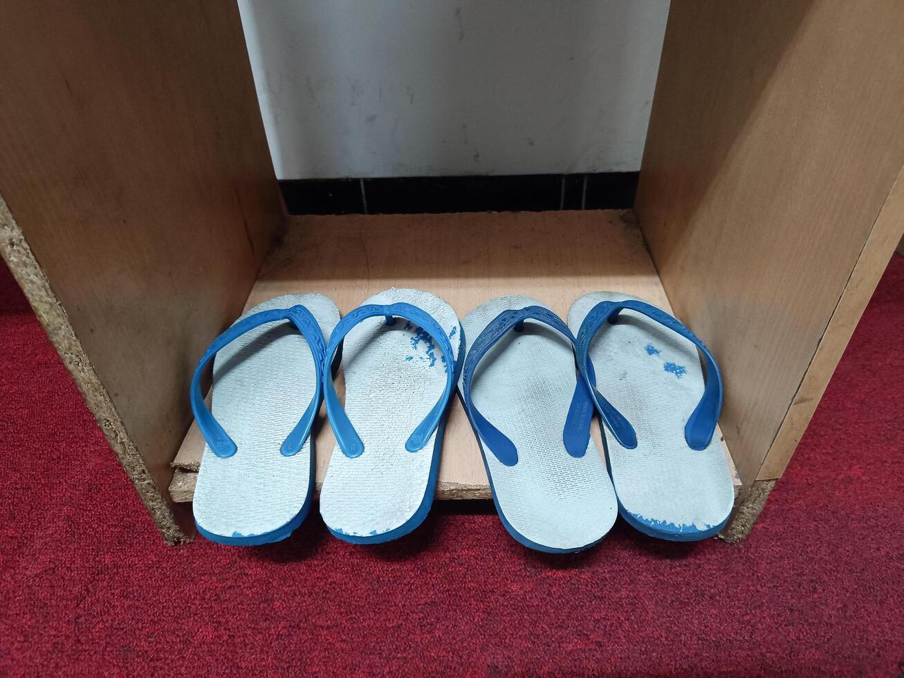 Two pairs of worn white flip-flops with blue straps, placed on a wooden shelf with a red carpet base. photo