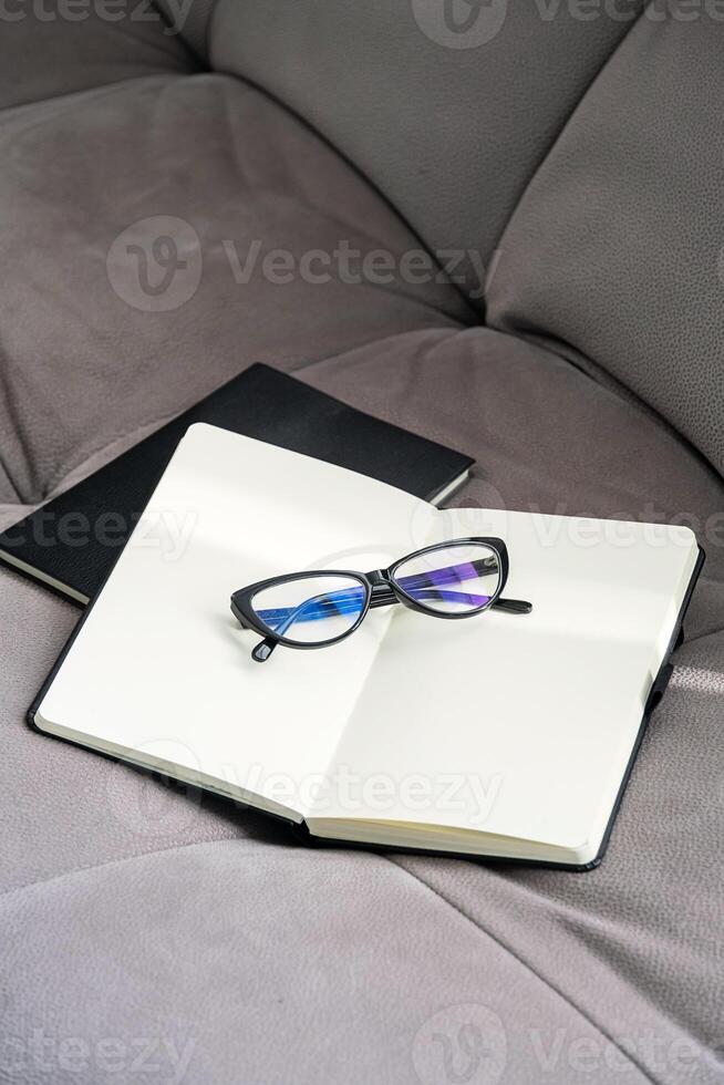 Pair of Stylish Black Glasses Rests on an Open Notebook Placed on Soft Gray Couch. photo