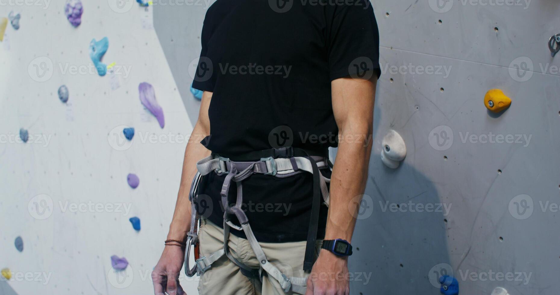 Young man of European appearance wearing safety belt, smiling looking straight into the camera, standing in front of a climbing wall in an indoor hall, dynamic shooting photo