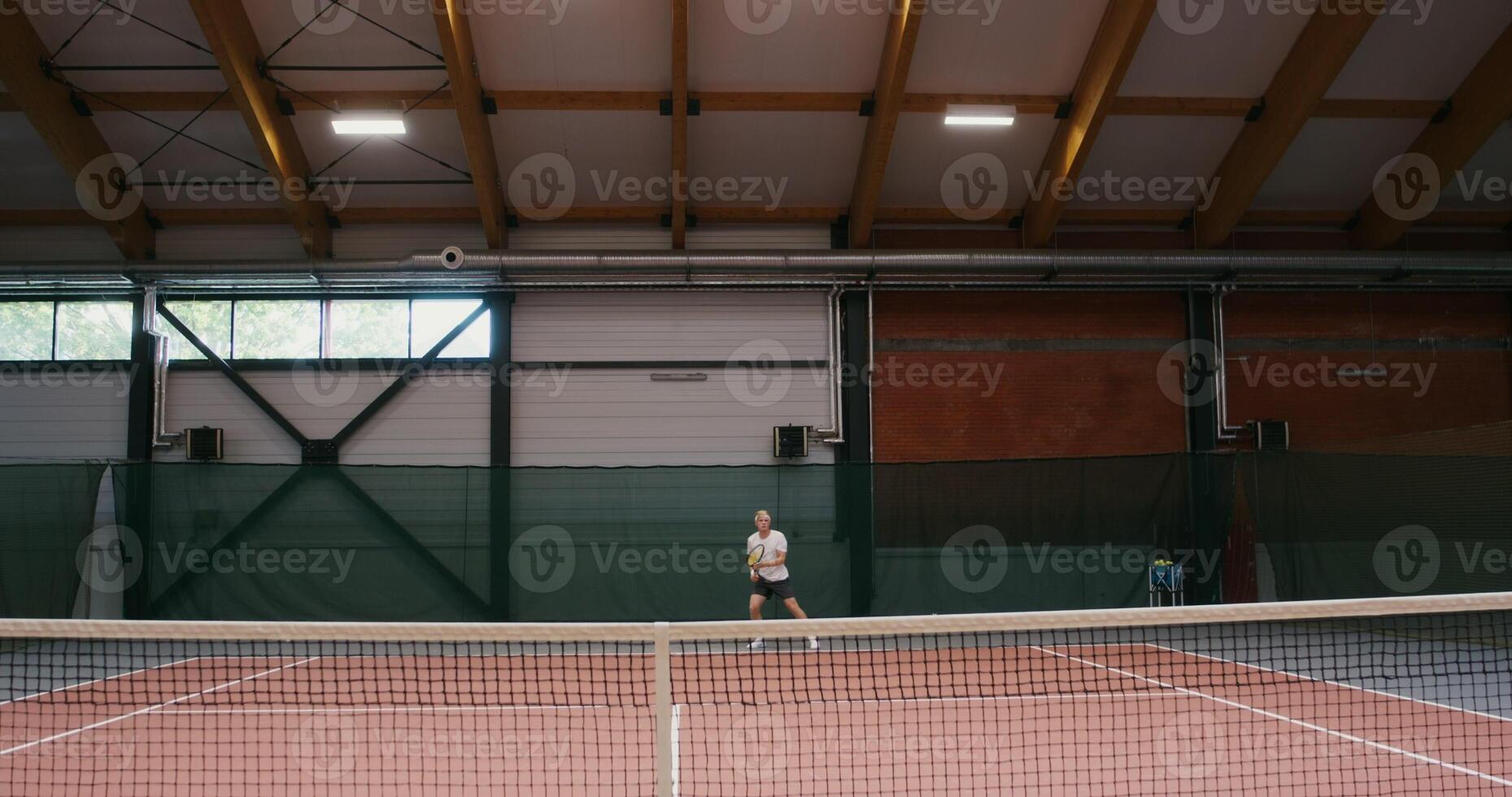 Young athlete plays tennis, bouncing and hitting a tennis ball with a racket throws it over the net. Competition in the indoor tennis court, front view photo