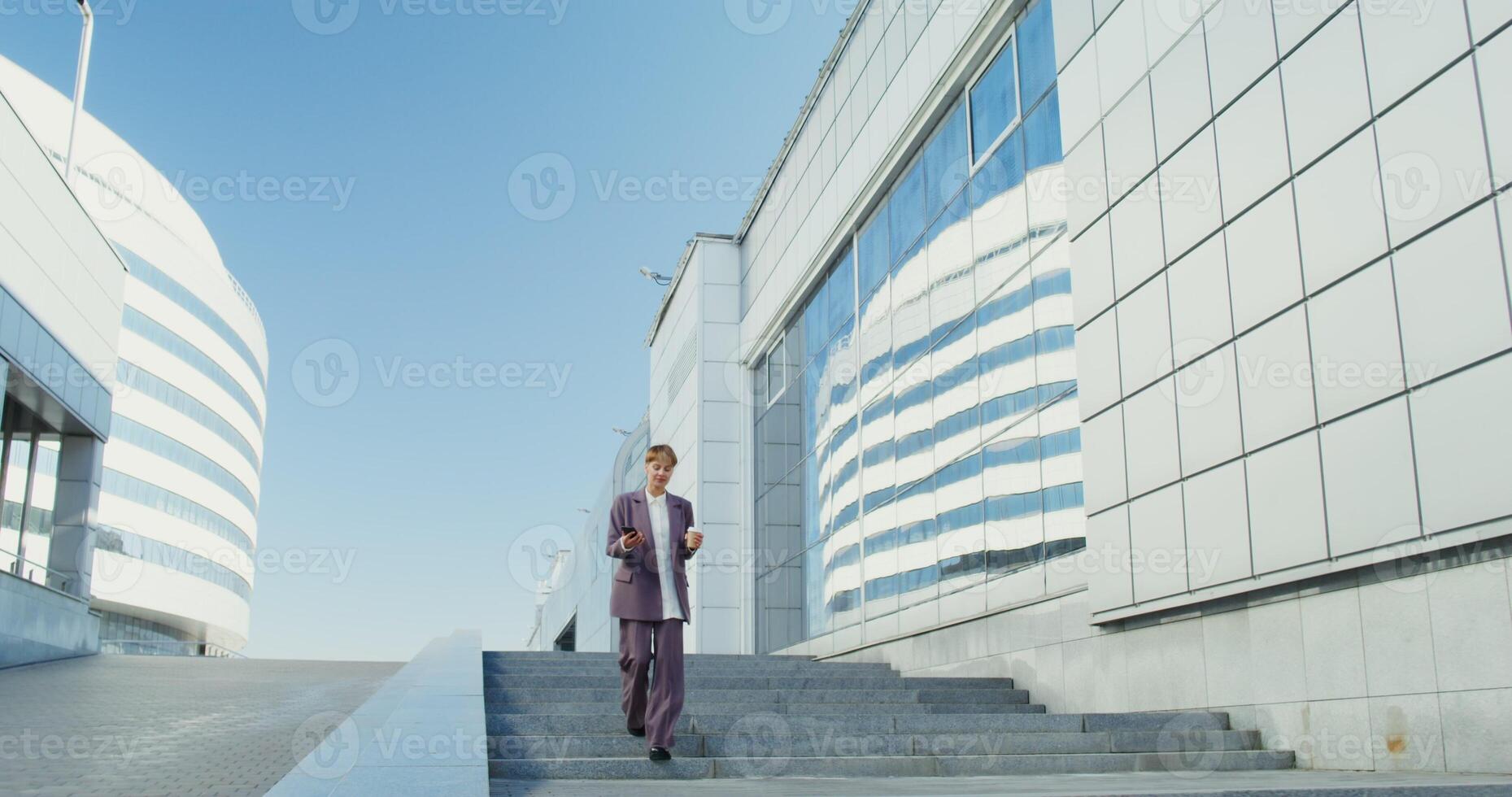 Woman of European appearance, dressed in a business suit, smiling while talking on a mobile phone and drinking coffee going down the street stairs between modern office buildings photo