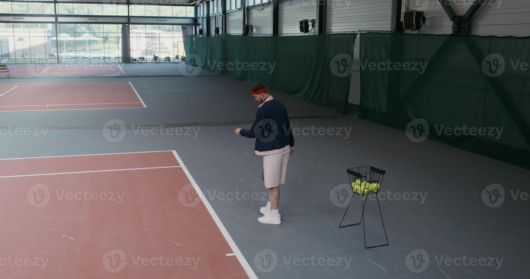 Man of European appearance takes a tennis ball from a basket full of balls and throws it over the tennis net, hitting the racket, playing in an empty indoor tennis court photo