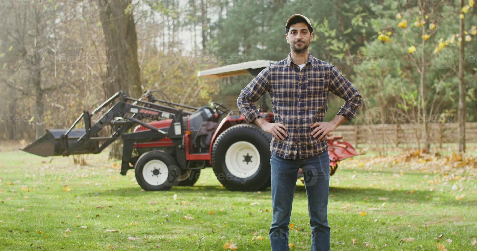 Young dark haired male farmer looking straight into the camera and folding his arms over his chest, as he stands near a large agricultural tractor outdoors photo