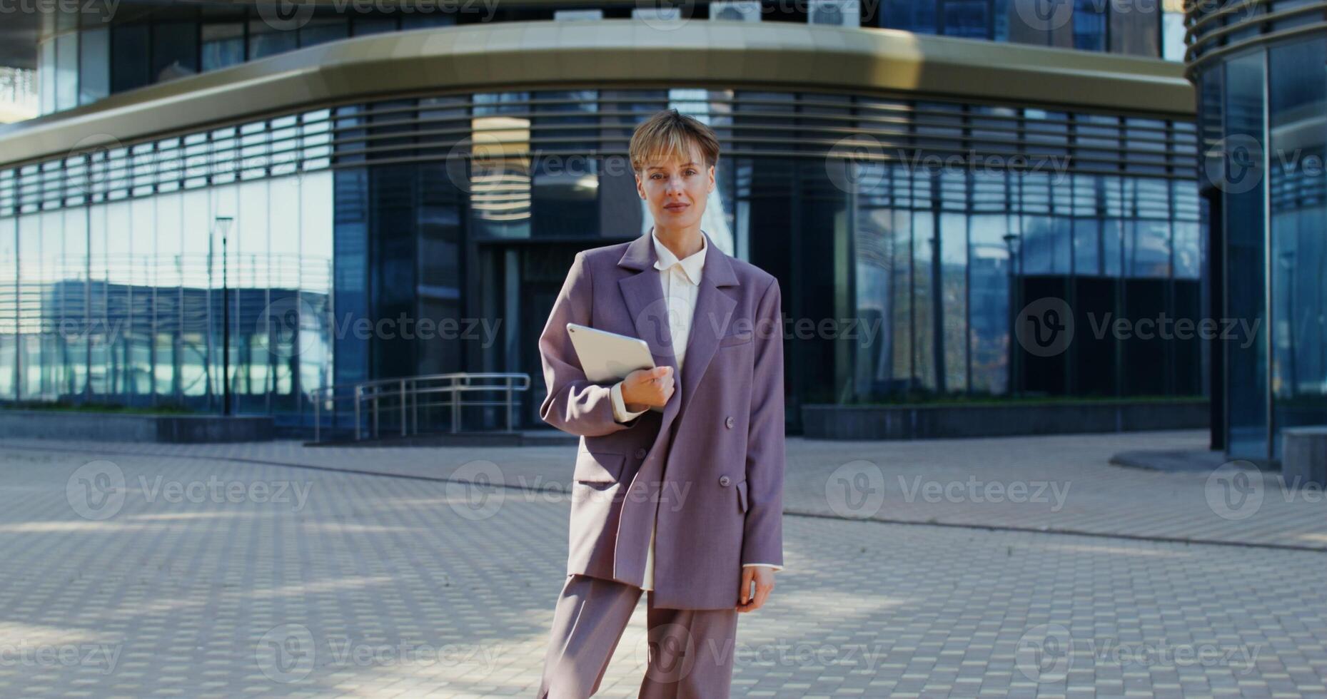 Young businesswoman of European appearance with a short haircut, dressed in a business suit, looks directly into camera while standing near a modern office building and holds a tablet in her hands photo