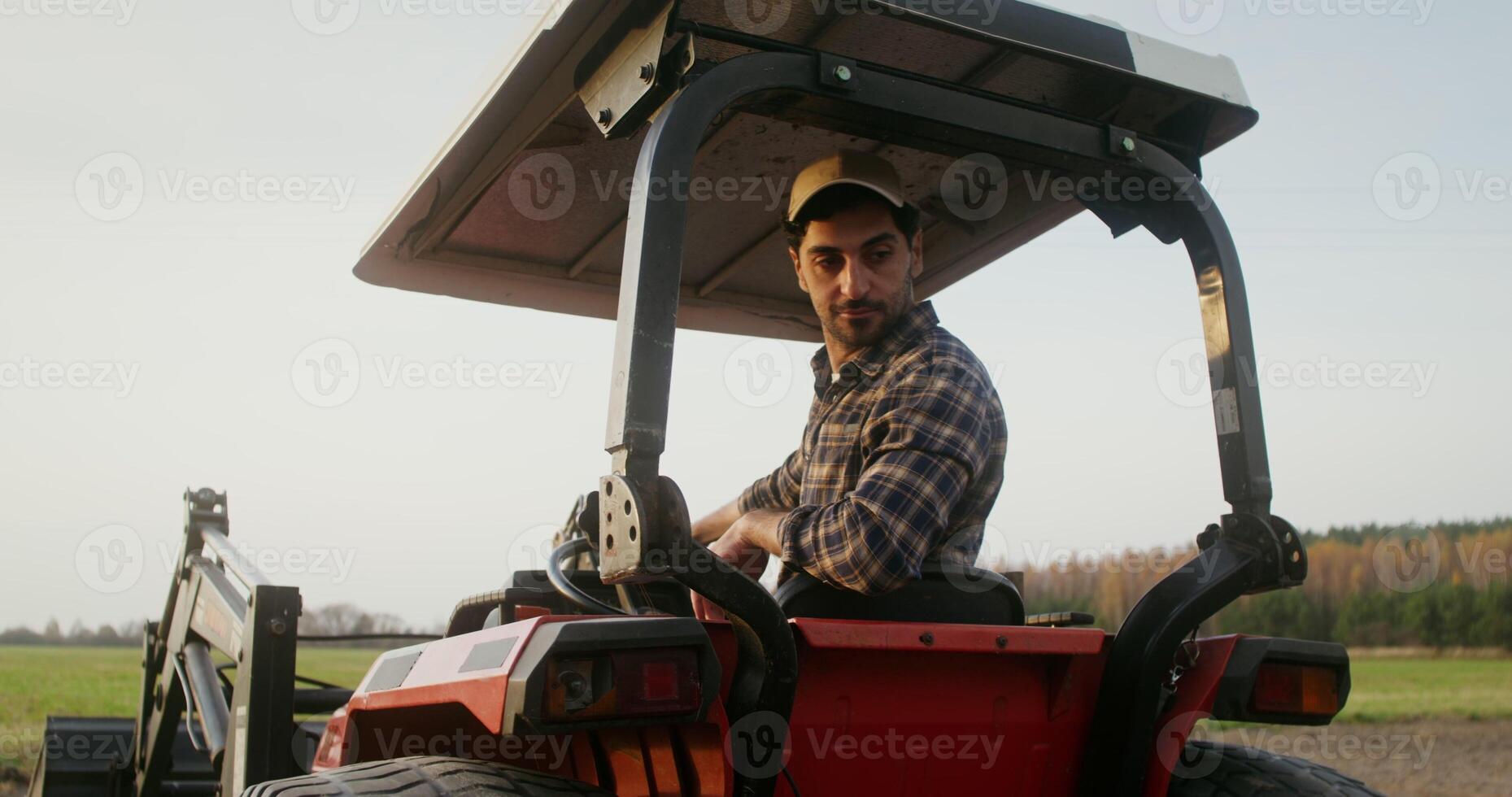 Young European farmer operates a farm tractor, while sitting in an open cab with canopy and turning back to control the evenness of the plowing strip photo