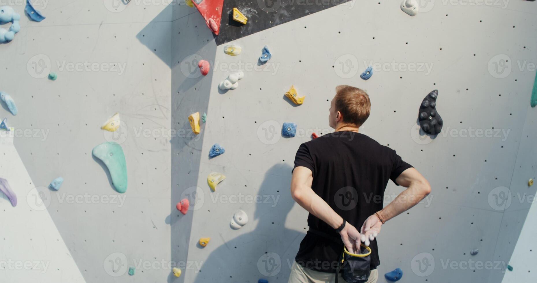Man of European appearance treats his hands with talcum powder, dropping them into a bag hanging on a belt behind his back, and begins to climb the climbing wall without belay. Filming from the back photo