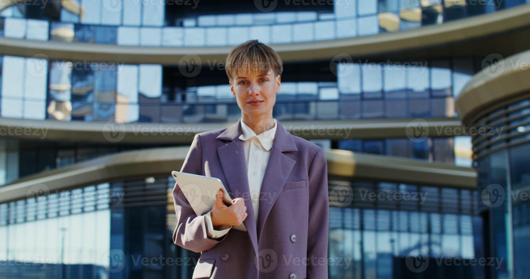Beautiful young european woman dressed in business, suit holding a tablet and smiling while looking at the camera while standing outdoors near a modern office building photo