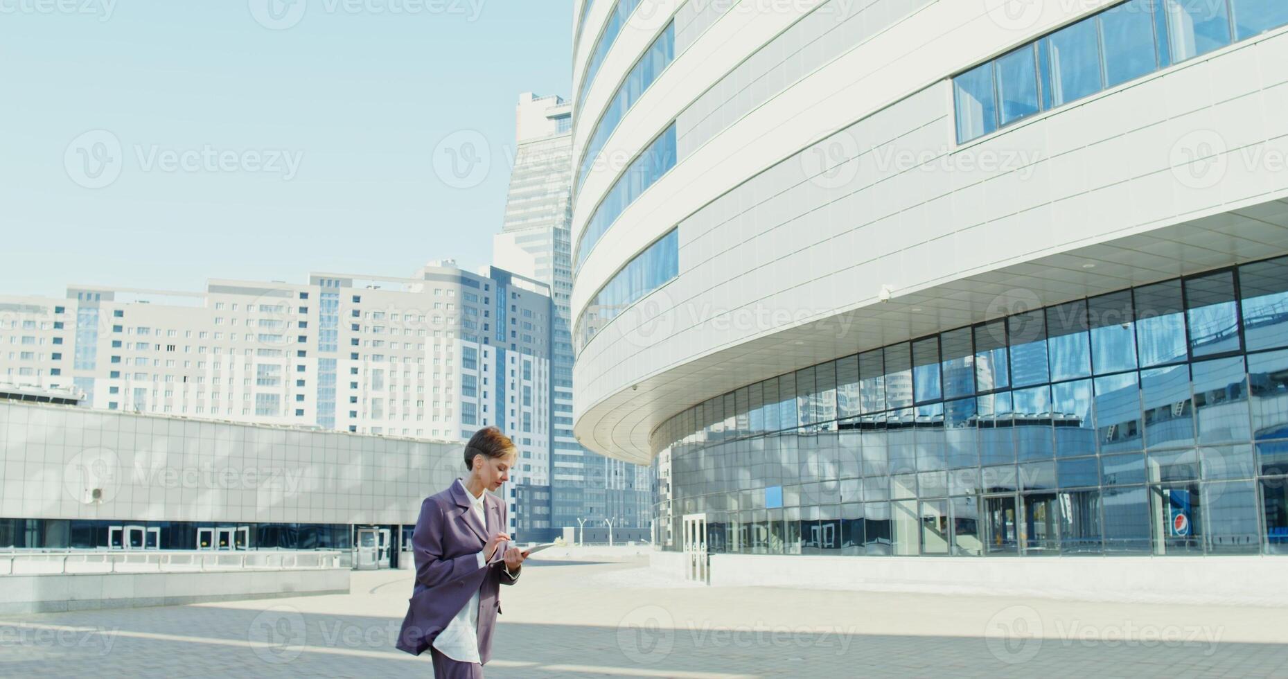 Young woman of European appearance with short hair, dressed in a man's business suit, smiling, walking around the city among modern buildings and using a tablet on the go photo