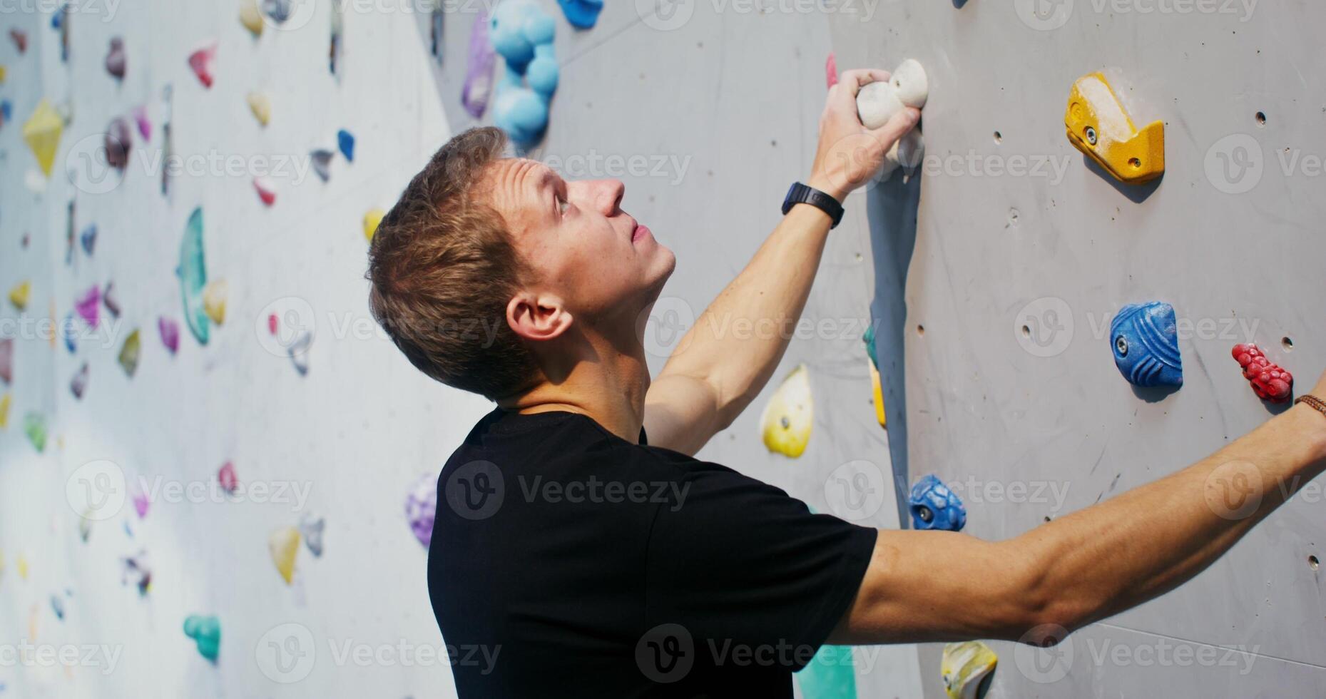 Man of European appearance, equipped with safety cables hanging from a strap, stands at the foot of the climbing wall and looks up, thinking over a route for climbing photo