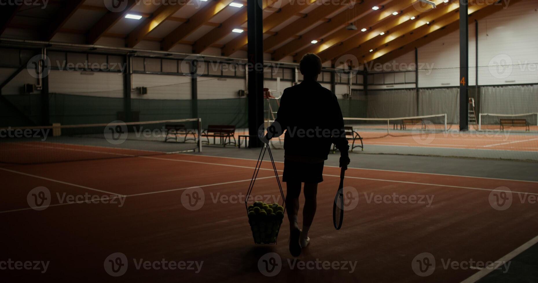 The lights turn on and young man of European appearance walks onto the tennis court, carrying a basket full of tennis balls, preparing for a training session alone. photo