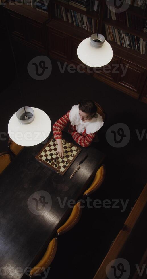 Top view of a young woman of European appearance plays a chess game alone, thinking long concentratedly between moves, sitting at a long table in the library, vertical photo