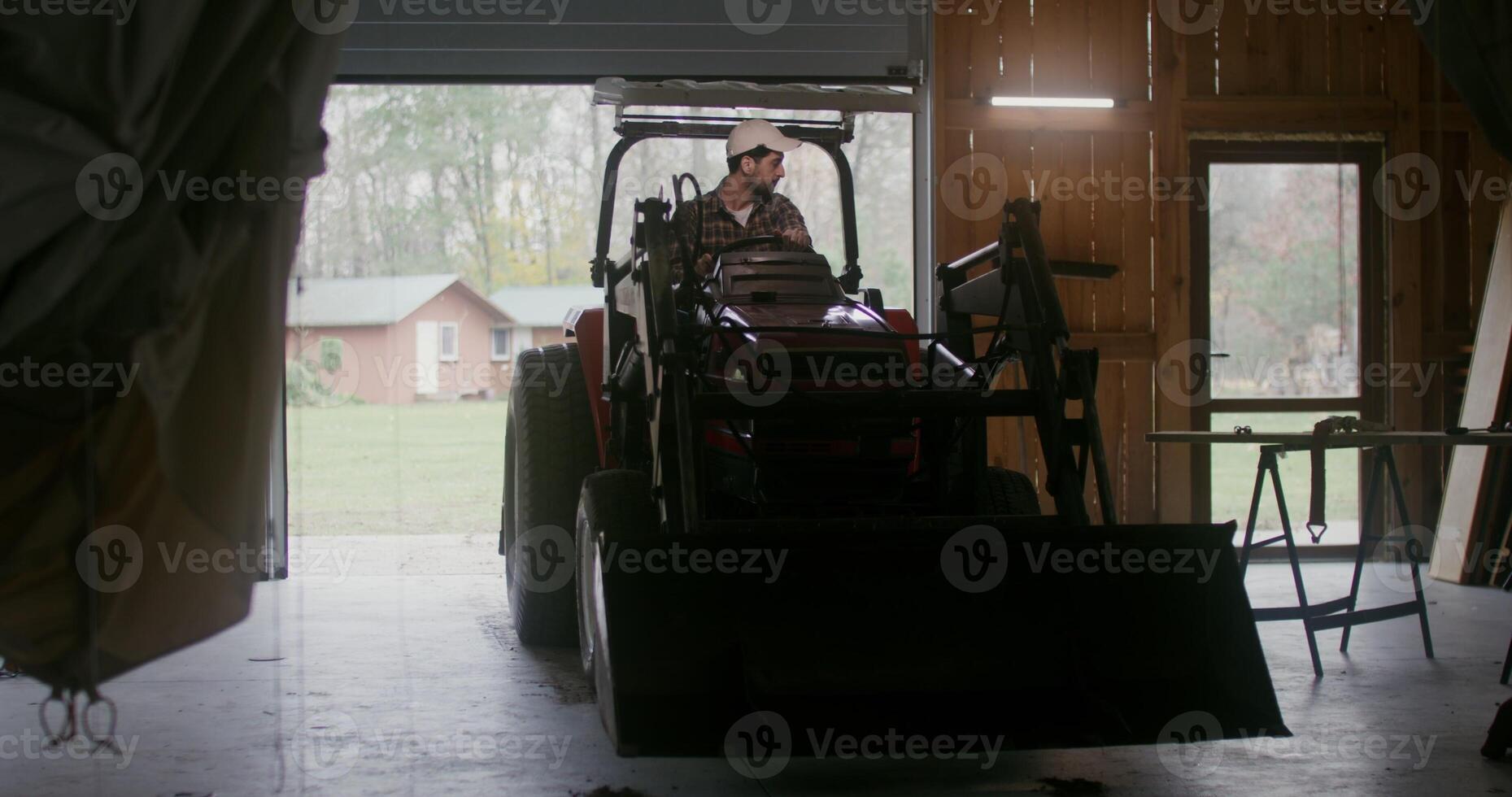 Young man of European appearance with a beard, starts a small agricultural tractor with a bucket and drives it out of the garage, backing it up, going to work on the farm on an autumn day photo
