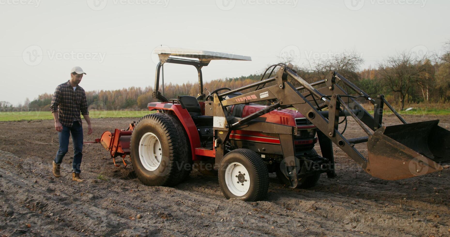 Young man of European appearance in a cap, walks through a plowed field to a tractor and sits on the seat of an open cabin behind the wheel photo