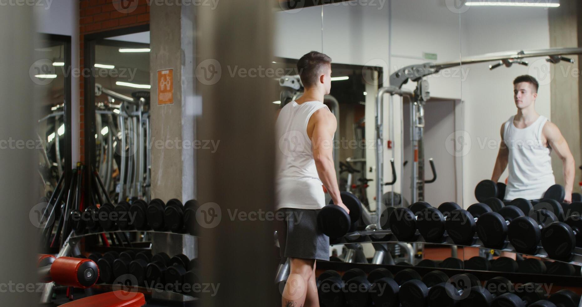 A man of European appearance, athletic physique starts workshop for biceps in a modern gym, standing in front of a mirror and choosing dumbbells photo