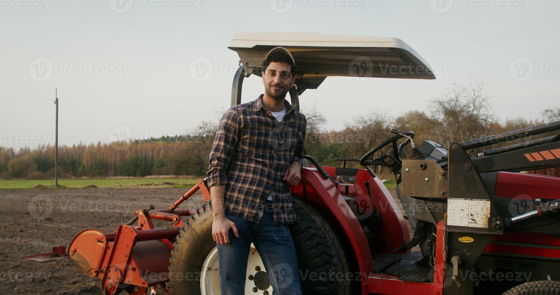 Young male tractor driver of European appearance stands leaning on an agricultural machine and smiles while looking directly into the camera, in a field on an autumn day photo