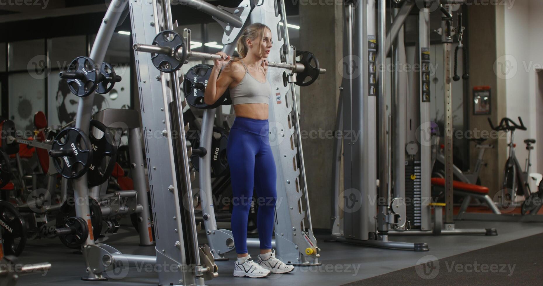 Young woman of athletic physique, dressed in a sports uniform, squats with a barbell on the exercise machine, doing in the gym. filming from the side, general plan of the gymnasium photo