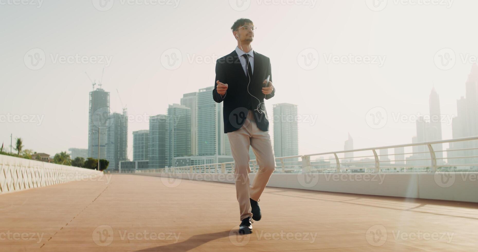 A man with glasses, wired headphones in his ears, dressed in a business suit dancing walking down the street with a mobile phone in his hands photo