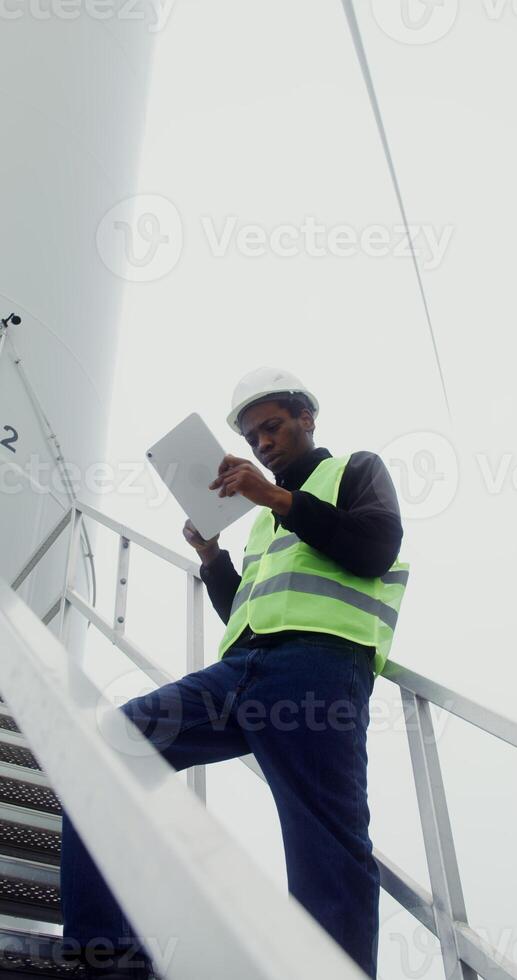A power plant worker in work gear uses a tablet with a serious look while standing on the steps near the entrance to an industrial wind generator, vertical , bottom view. in 4k, red komodo photo