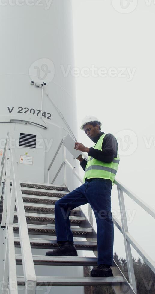 A power plant worker in a green vest and hard hat uses a tablet with a serious look while standing on the steps near the entrance to industrial wind generator, vertical . in 4k, red komodo photo
