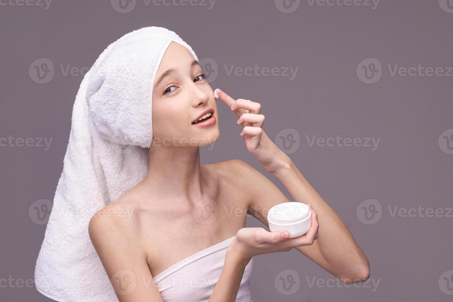 Young caucasian female applying skincare cream with towel wrapped hair on gray background. photo