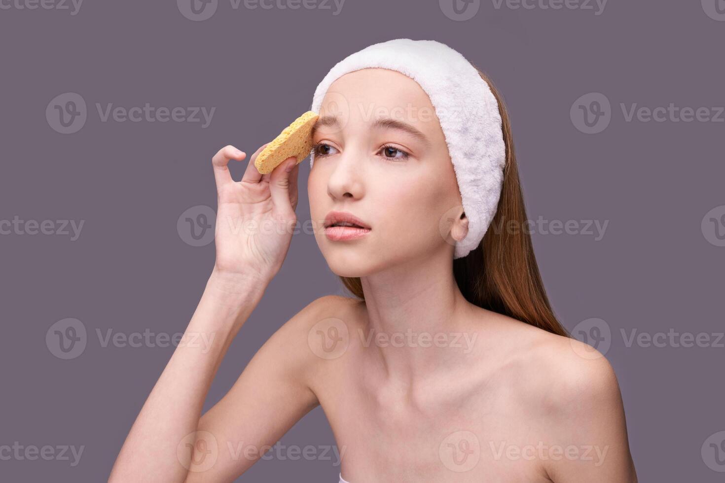 Young caucasian female with headband cleansing face using natural sponge on purple background. photo