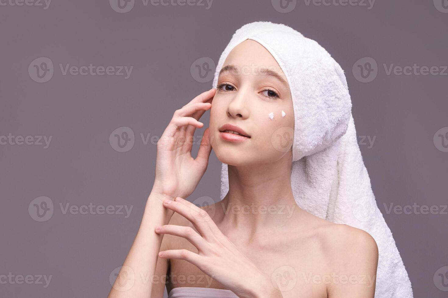 Young caucasian female applying skincare cream with towel on head in minimalist setting. photo
