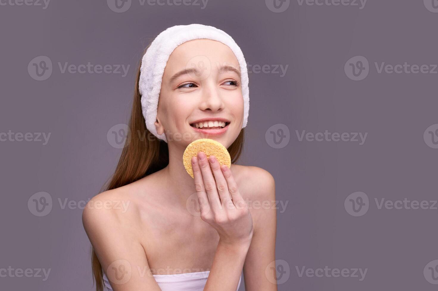 Smiling caucasian female teen using facial sponge for skin care routine with headband on grey background. photo