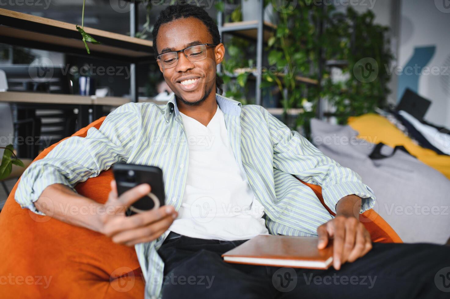 Smiling university student using smartphone and holding book in modern library photo