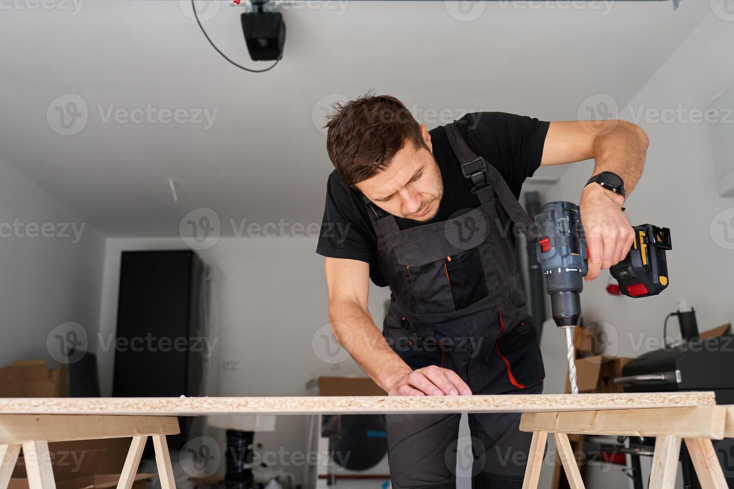 Man in work overalls bib using cordless power drill to make hole in laminated wood board. Male carpenter working in home workshop photo