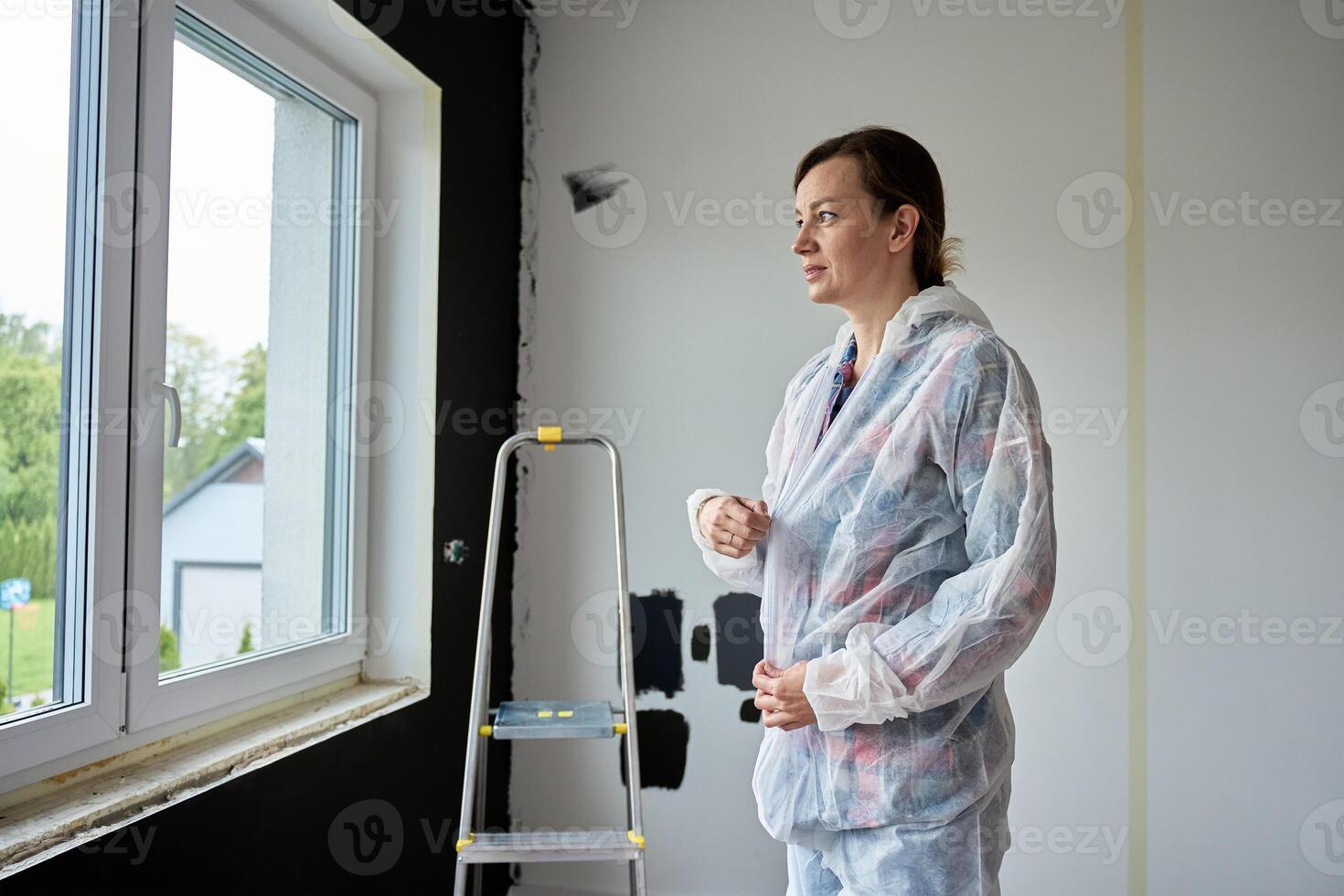 Woman in protective coveralls standing in front of unpainted wall with paint samples. Person preparing to paint interior wall at home. Concept of home renovation and interior upgrade photo