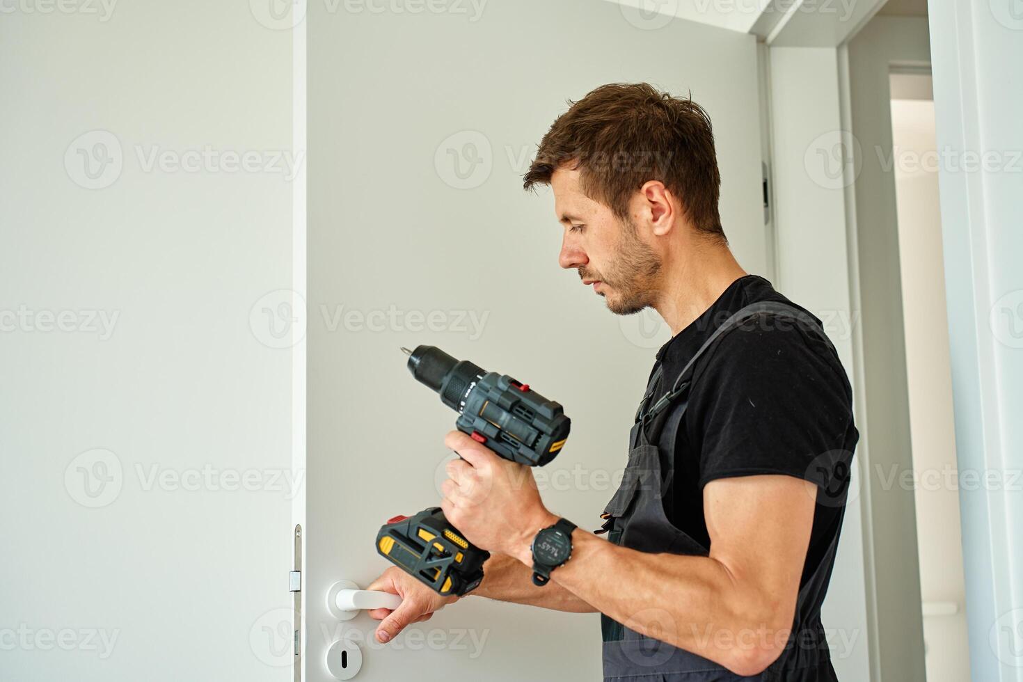 Man holds cordless drill and installs white door handle in modern interior. Handyman fitting door in apartment, using screwdrive photo