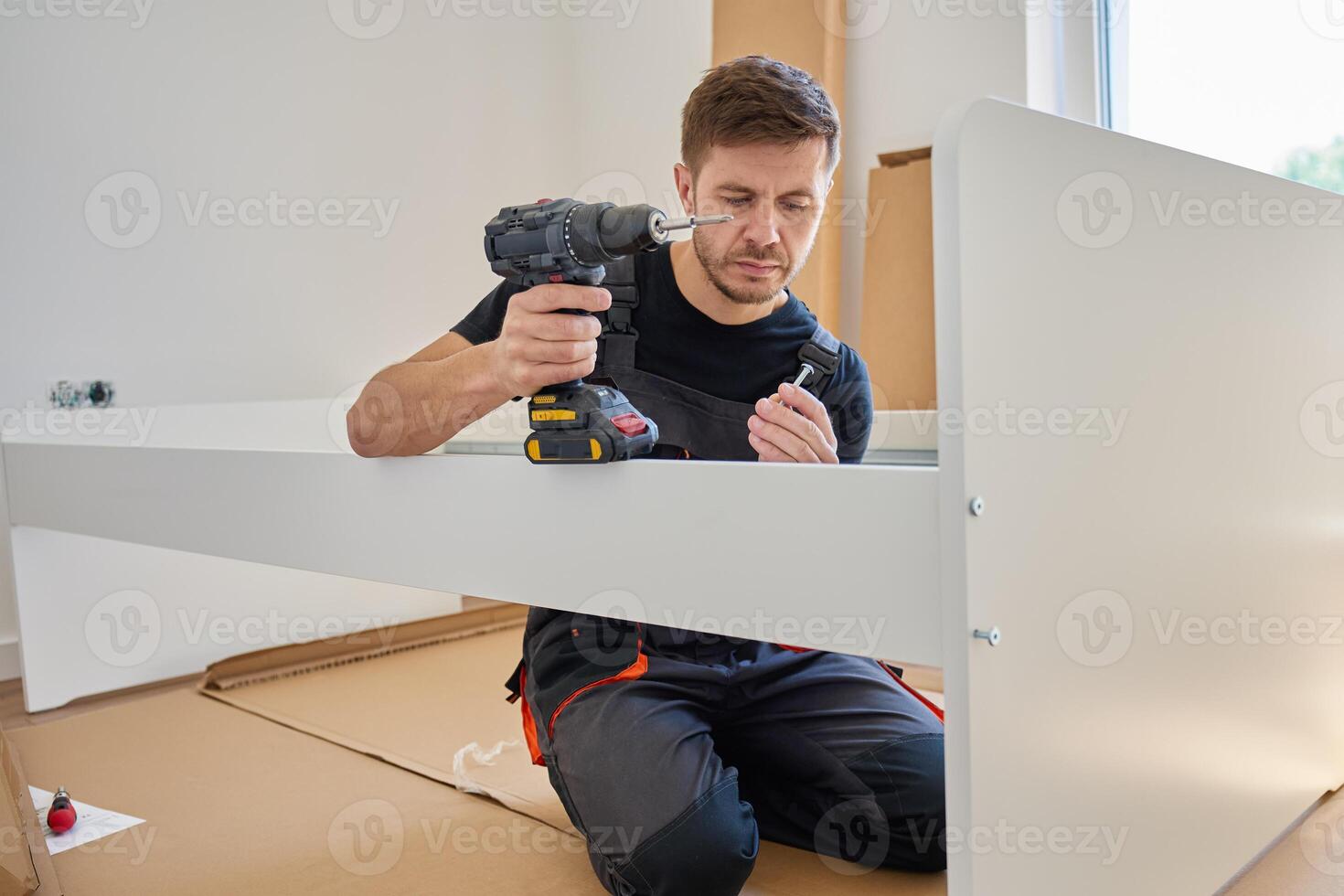Focused man in bib overalls connects details of new bed in bedroom using electric drill. Assembling furniture at home photo
