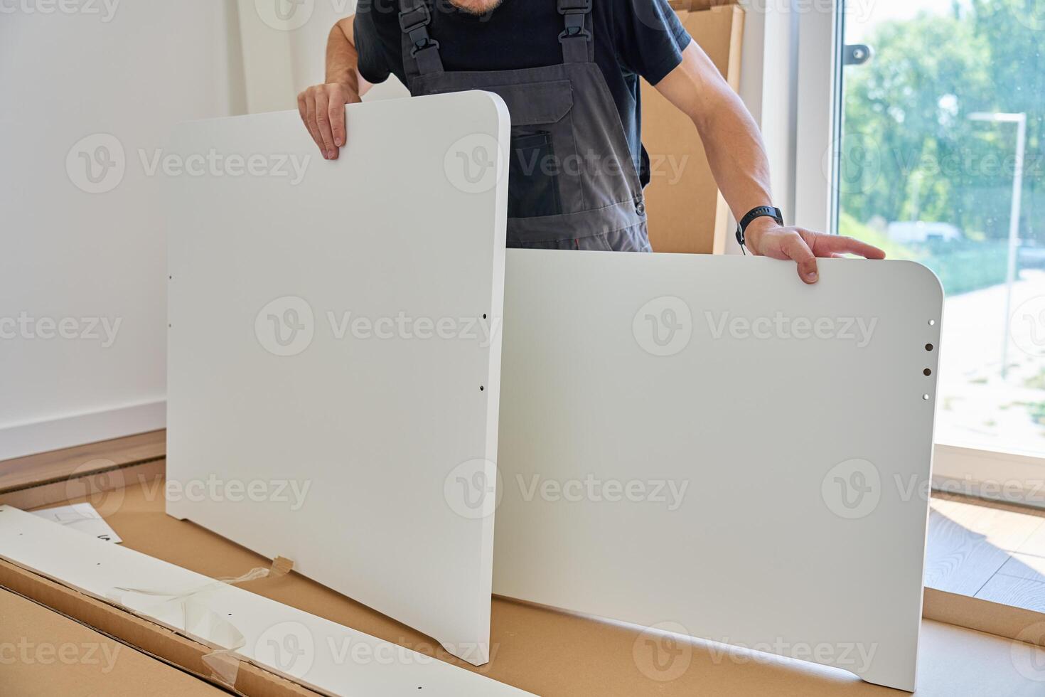 Man in work overalls assembles white furniture panel near unpacked parts and tools at home. Handyman preparing part of bed for assembly in bedroom photo