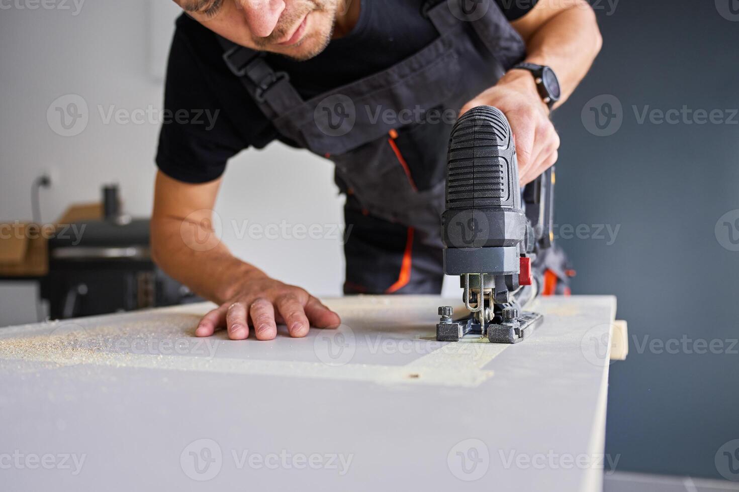 Worker in workwear using handheld cordless electric jigsaw to cut wooden board in garage photo