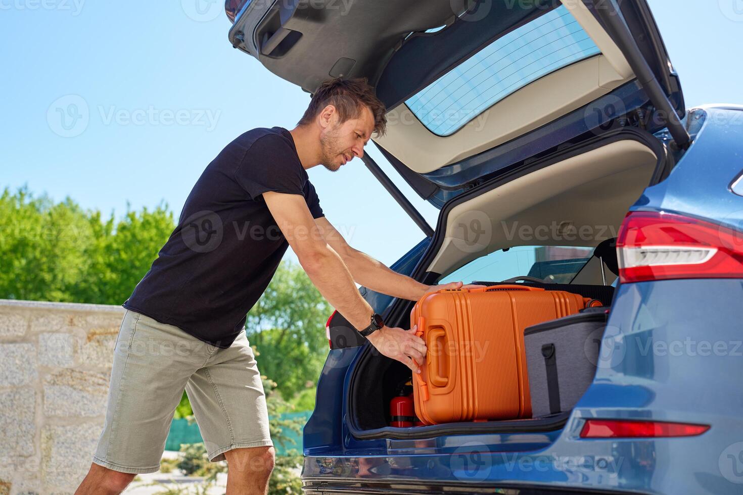 Man loading orange suitcase into car trunk on sunny day. Concept of preparing for summer travel, vacation or road trip with luggage photo