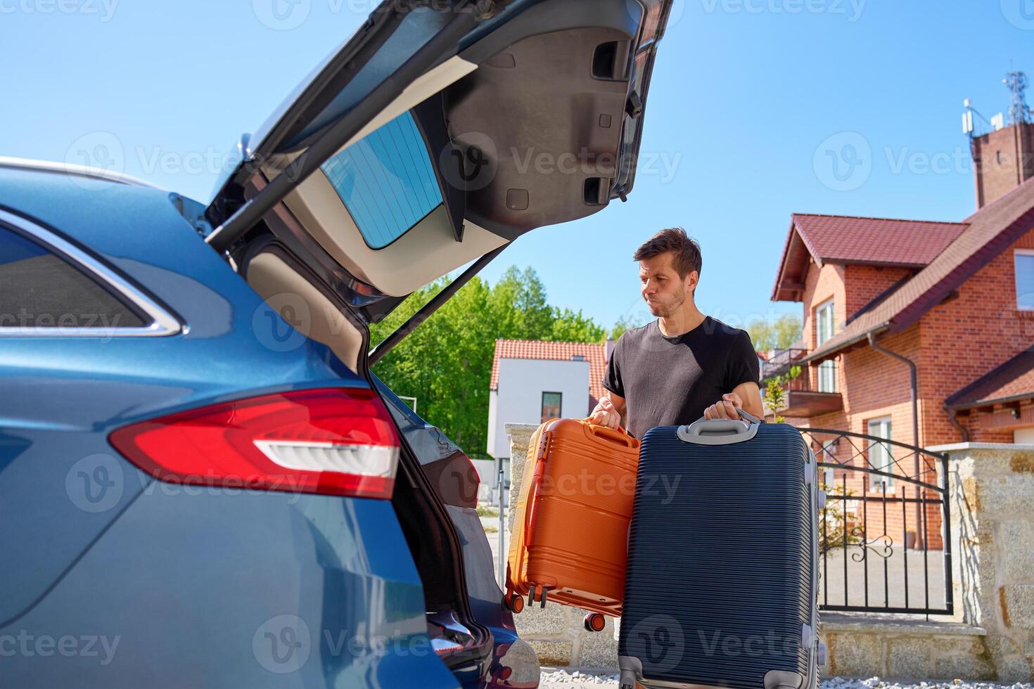 Man holding orange and blue suitcases near parked car in front of suburban brick house. Loading luggage in vehicle. Concept of moving day, relocation or travel preparation photo