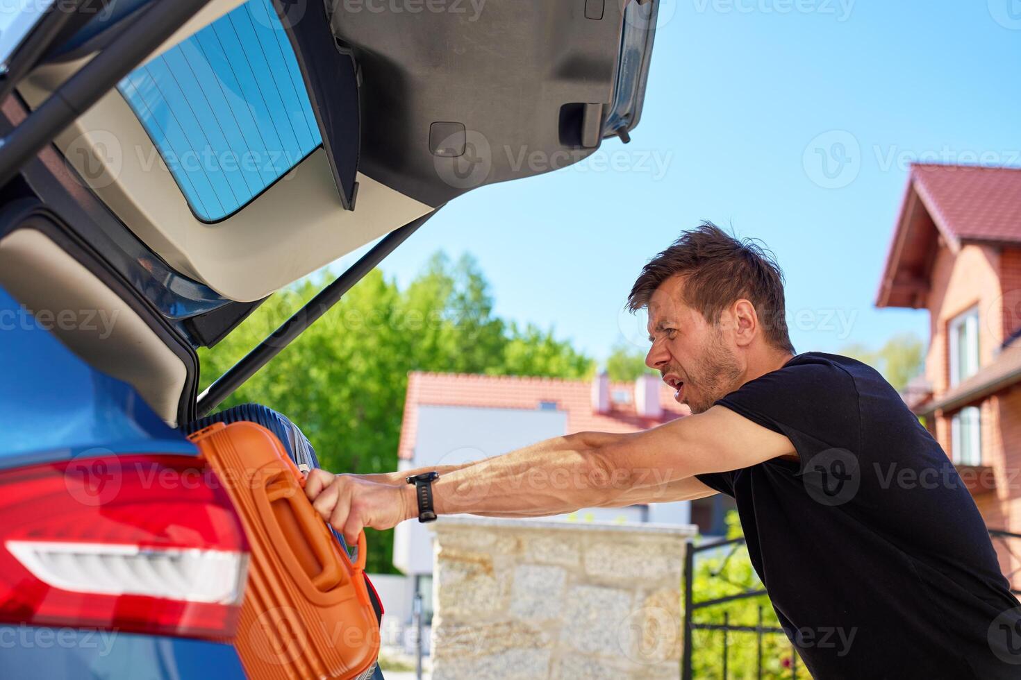 Man pushing orange suitcase in full car trunk near suburban house. Loading luggage in vehicle. Concept of moving day, relocation or travel preparation photo