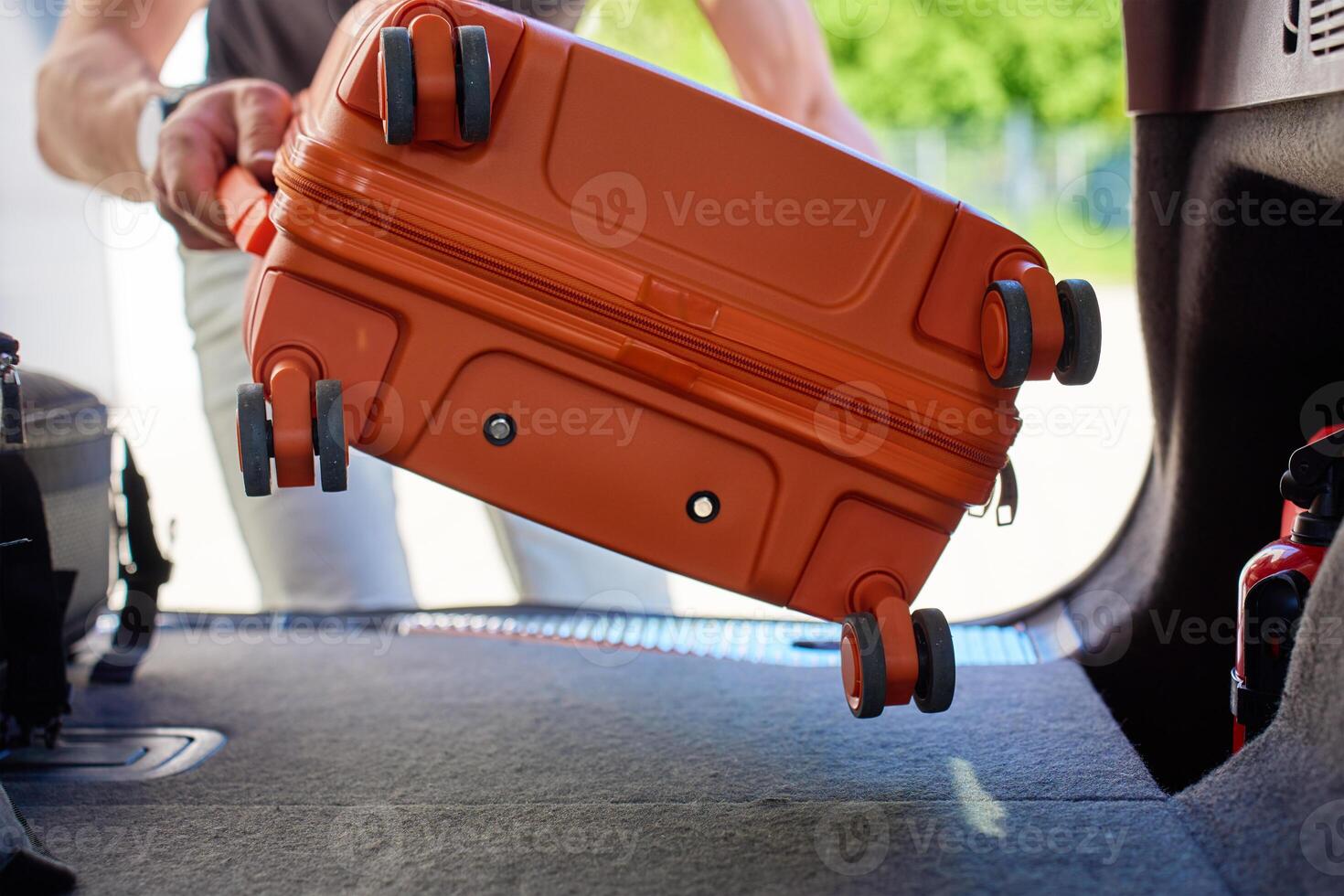 Close up of man loading orange suitcase into open car trunk outside residential house. Inside view of male traveler placing luggage in vehicle before road trip. Preparation before travel or departure photo