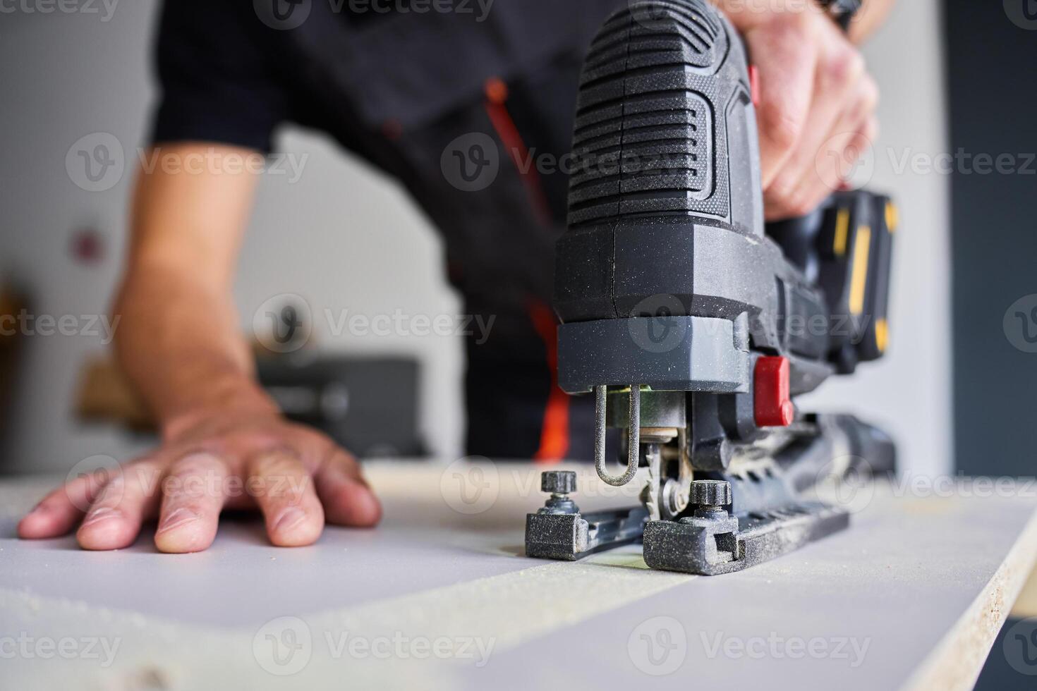 Worker in workwear using handheld cordless electric jigsaw to cut wooden board in garage. Skilled carpenter working in workshop photo