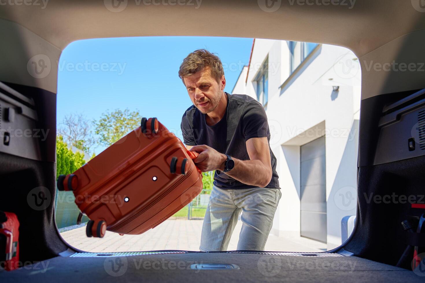 Man placing orange rolling suitcase into open vehicle trunk, preparing for journey or vacation on sunny day. Inside view of traveler loading luggage in car. Travel or weekend road trip concept photo