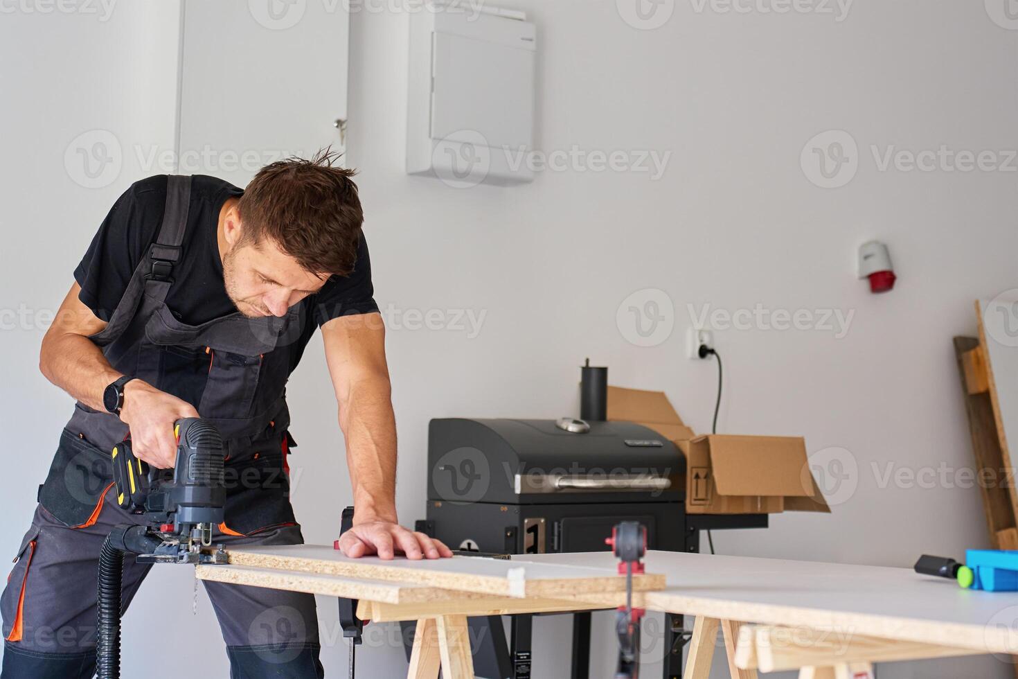Carpenter cuts wooden board with electric jigsaw in home workshop. Man in workwear working with wood on workbench in garage. Woodworking and carpentry concept photo