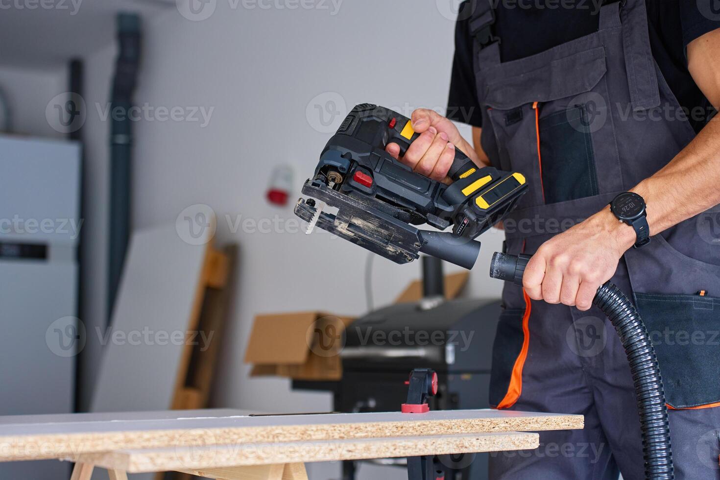 Man in workwear attaching vacuum hose to electric jigsaw for woodworking on workbench in home workshop photo