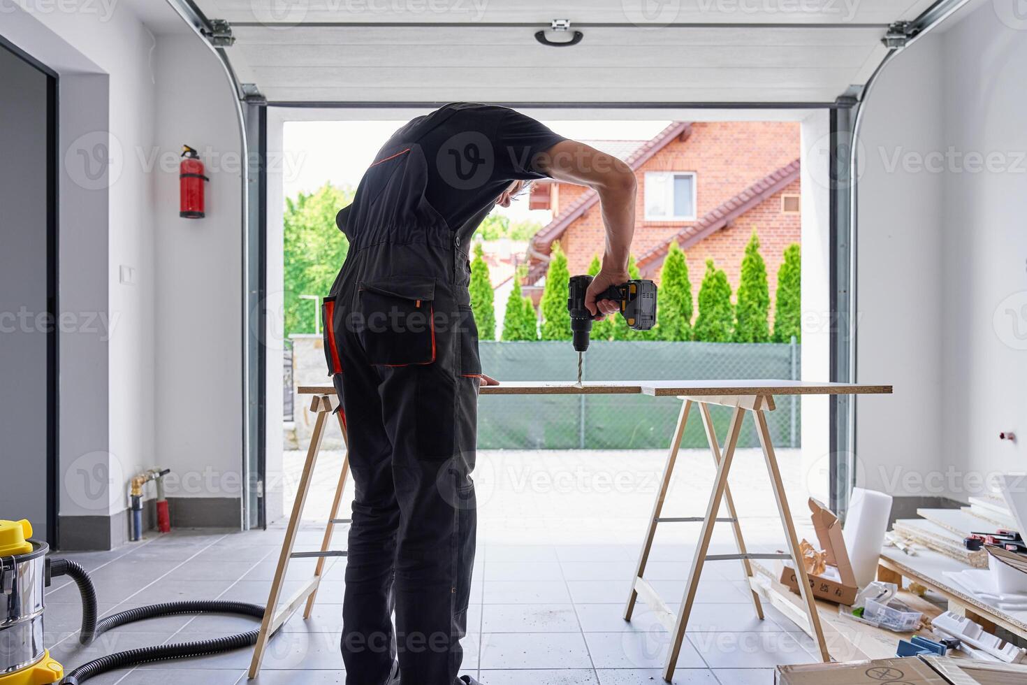 Worker using power drill on wooden board supported by sawhorses in home garage workshop. Man working in workshop, drilling hole in chipboard for furniture assembly photo