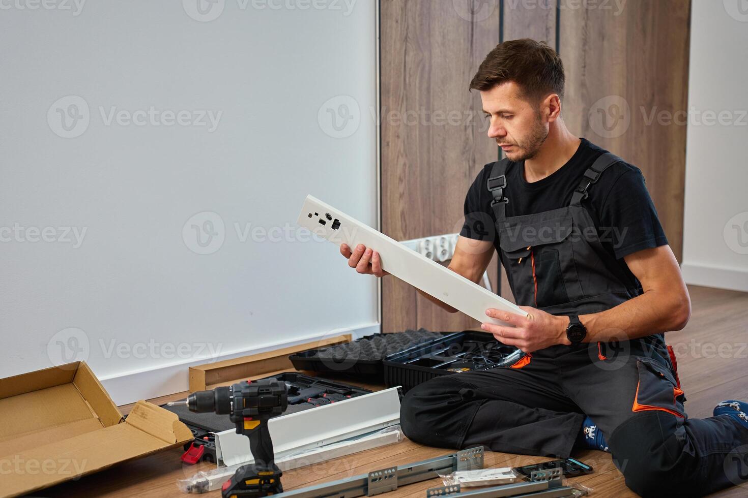 Man in overalls assembles furniture, examining white panel and surrounded by tools. Handyman in work clothes preparing parts pf cabinet to assembly. DIY furniture project photo