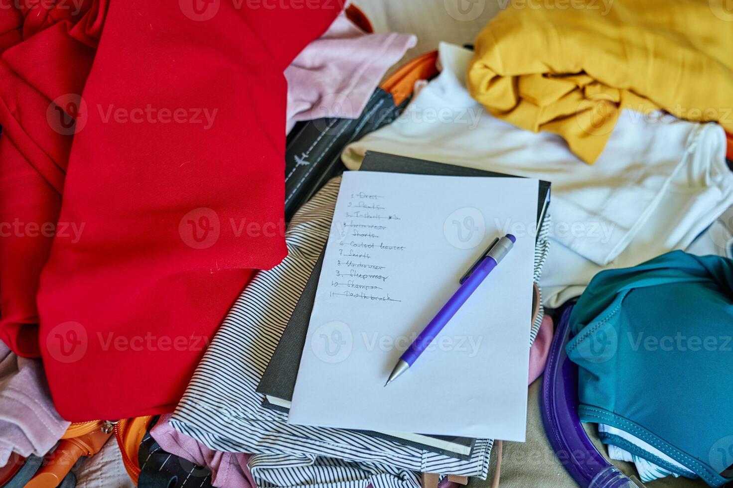Handwritten checklist and pen on top of colorful folded clothes inside open suitcase. Concept of travel planning and packing essentials for trip photo