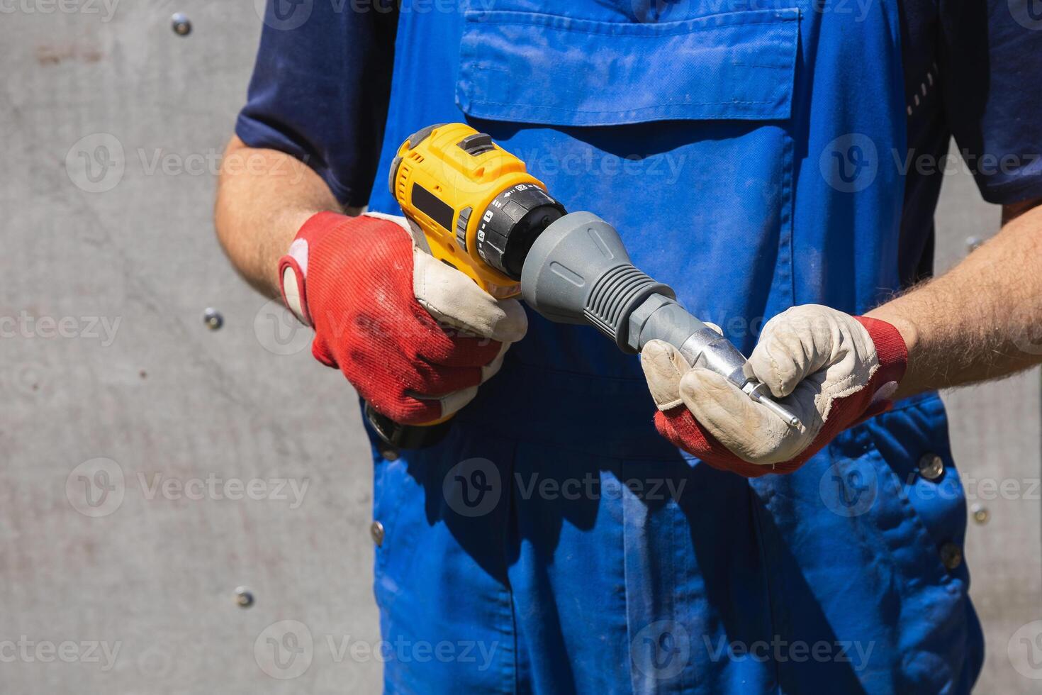 A worker in a blue jumpsuit holds an electric screwdriver with a riveter attachment and an exhaust rivet against a slate wall photo
