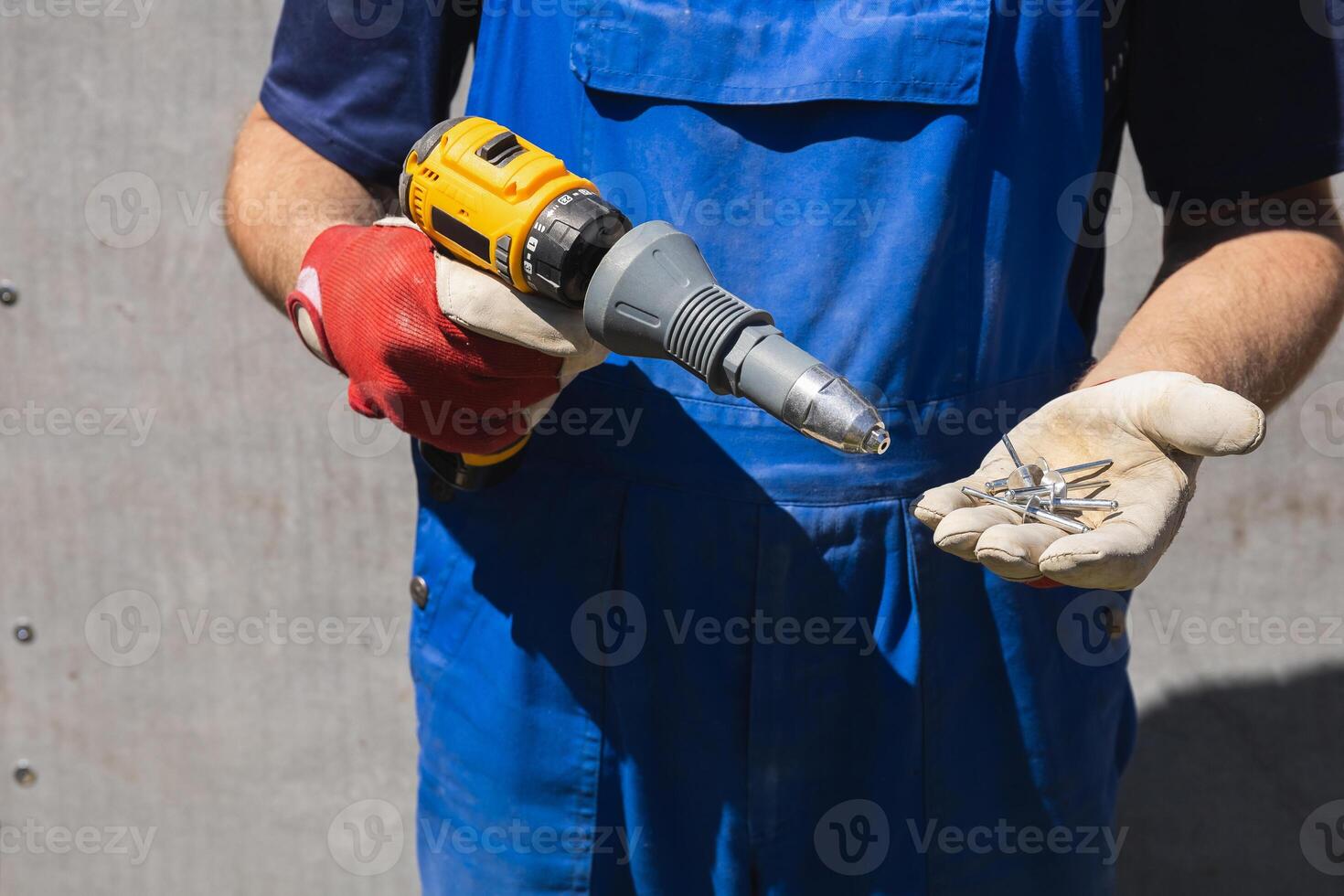 A man in a blue jumpsuit holds electric screwdriver with a riveter attachment and exhaust construction rivets on the background of a slate wall photo