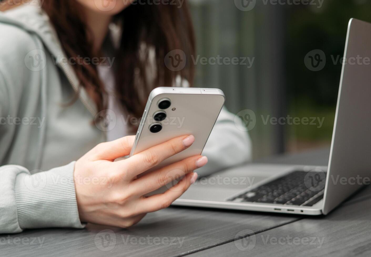 A person using a smartphone while sitting with a laptop on a wooden table outdoors, symbolizing multitasking and modern technology. photo