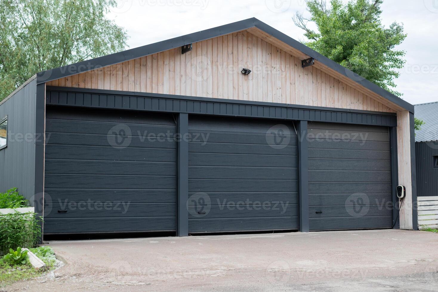 Modern three-door garage building with black automatic gates and light wood facade, located on a clean concrete driveway. photo