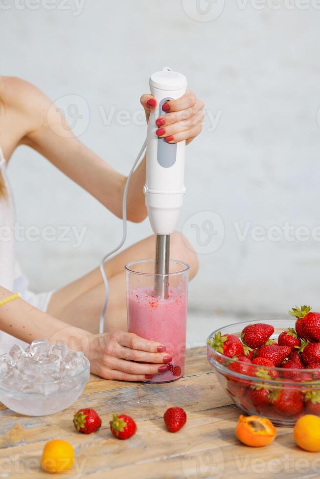 Woman using hand blender to mix fresh strawberry smoothie in glass on wooden table with fruits and ice. Healthy summer drink preparation, close-up hands and ingredients. photo